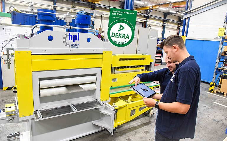 Zwei Mitarbeitende der hpl-Neugnadenfelder Maschinenfabrik GmbH besprechen den digitalen Entwurf einer Sondermaschine vor einer hpl-Anlage. Das DEKRA-Siegel verweist auf die erfolgreiche ISO 9001-Zertifizierung. 